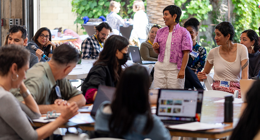 An individual in a pink shirt addresses a crowd in a cafe setting