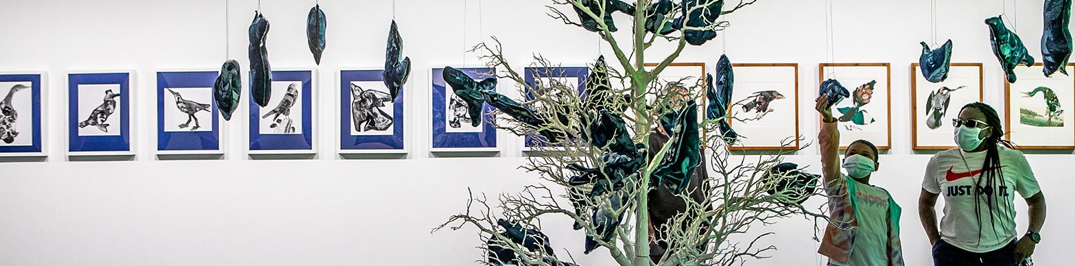 Two Individuals in masks stand in a gallery space observing an artificial tree