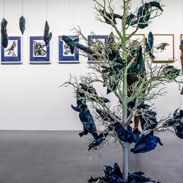 Two Individuals in masks stand in a gallery space observing an artificial tree
